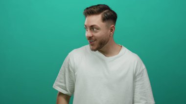 Beyaz tişörtlü, sakallı genç adam yeşil arka planda tek başına duruyor. Stüdyo ortamında tuhaf bir ifadeyle yan tarafa bakıyor.