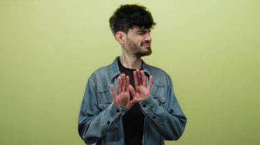 Tereddüt ve belirsizliği vurgulayan, parlak sarı bir arkaplan önünde kot ceket giyen, şüpheci bir ifadeyle isteksizliğini ifade eden genç bir adam..