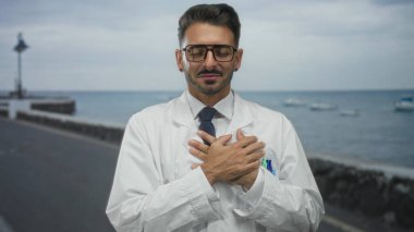 Laboratuvar önlüklü genç İspanyol bilim adamı her iki elini göğsünün üzerinde tutuyor ve gün ışığında deniz kenarında bir sokakta gülümsüyor; minnettarlık.