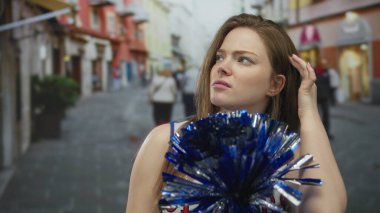 Amigo kız, şehrin kalabalık caddesinde elinde metalik mavi ponponu tutarken saç ayarlıyor. Kendinden şüphe ediyor..