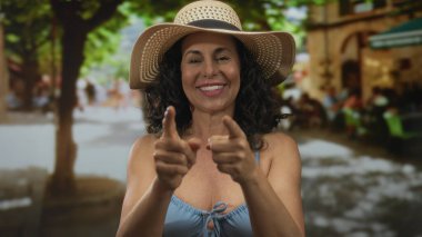 Mavi bir bikini ve hasır şapkayla gülümseyen kadın güneşli bir sokakta insanların ve kafelerin bulanık geçmişiyle, açık hava neşesi ve yaz rahatlığının özünü yakalıyor..