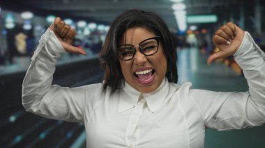 Tren istasyonunda kendinden emin bir şekilde gülümseyen, kendini gösteren başparmaklarıyla işaret eden, gözlük ve beyaz gömlek takan, kapalı alanda kendini ifade etme gücünü yakalayan bir kadın..