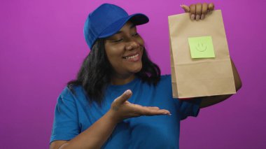 Mavi şapkalı kurye kadın elinde kese kâğıdıyla mor stüdyoda avucunun içini sunar. Mutluluk..