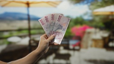Açık hava restoranının terasında elinde Türk lirası banknotları olan bir adam güneşli gökyüzünün altındaki bir caddede kahve içiyor..