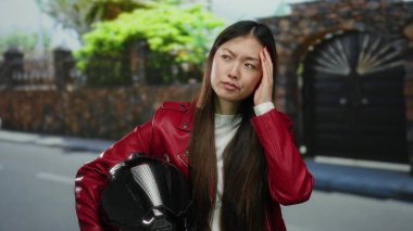 Şehir sokağında miğfer tutan kırmızı ceketli kadın, taş duvarlı ve kapılı bulanık şehir zeminine karşı düşünceli ve düşünceli görünüyor..