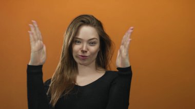 Genç gülümseyen beyaz kadın, stüdyoda iki avucunu da turuncu duvarla kafasının yanına kaldırıyor; neşeli açıklık.
