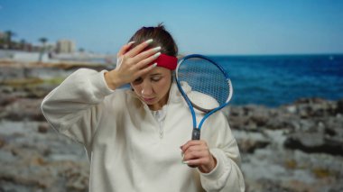 Sahilde tenis raketi tutan bir kadın, okyanus manzarası ve günlük kıyafetlerle düşünceli bir görünüm sergiliyor..