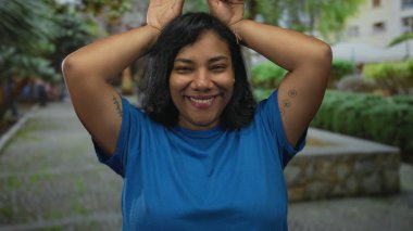 Gülümseyen ve kulakları olan bir kadın, yapraklı bir parkta elleri başının üzerinde mavi bir gömlek giyerek hareket eder..