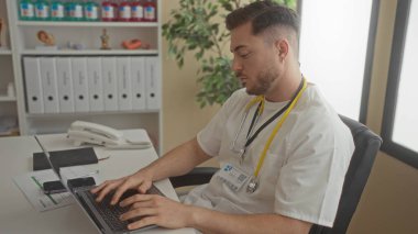 Steteskop takan sakallı genç bir adam klinikte dizüstü bilgisayarda çalışıyor. Arkasında dosyalar ve tıbbi ekipmanlar var..