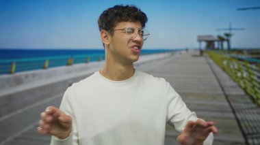 Gözlüklü bir adam beyaz bir süveter giyer manzaralı bir caddede parmağını gösterir; neşeli bir heyecan.