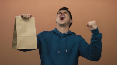 Genç İspanyol adam, izole edilmiş kahverengi duvar arkaplanına kağıt torba dayayarak heyecanını kutluyor..