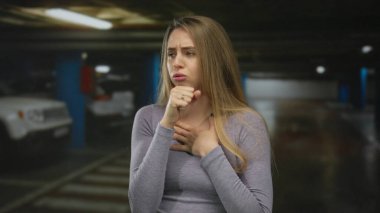 Kapalı bir otoparkta arka planda arabalarla öksüren, günlük gri bir kıyafet giyen, sıkıntı ya da hastalık belirtileri gösteren bir kadın..