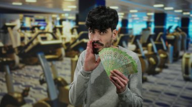 Spor salonunda spor aletleriyle Rus rublesi taşıyan bir adam, kapalı bir spor salonunda güvenle gülümsüyor..