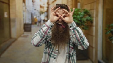 Sakallı adam iki eliyle parlak bir işaret yapar ve eski bir şehir sokağında dilini dışarı çıkarır..