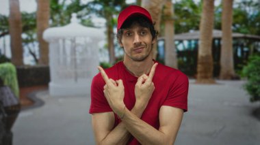 Kırmızı şapkalı ve kırmızı gömlekli adam caddede çapraz kollarını gösteren palmiye ağaçları ve banklar; belirsizlik şüphesi şaşkınlık şaşkınlık şaşkınlık şaşkınlık şaşkınlık şaşkınlık şaşkın.