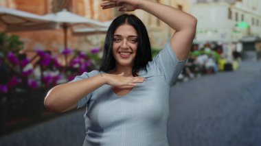 Canlı bir şehir caddesinde neşeli bir şekilde el kol hareketi yapan kadın, şehir manzarasının ortasında sıcak bir gülümsemeyle vücut özgüveni ve pozitifliğini gösteriyor..