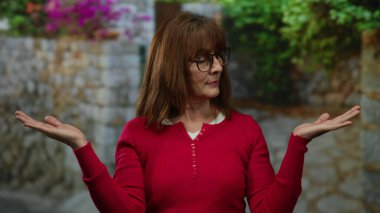 Kırmızı kazaklı, gözlüklü bir kadın sakince poz veriyor şehir sokağında bitkilerle, sükunet ve sükuneti temsil ediyor..