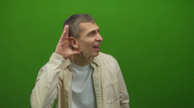 Canlı yeşil duvara karşı el ele tutuşarak dinleyen, izole bir ortamda merak ve konsantrasyon taşıyan, günlük beyaz gömlek giyen, nişanı vurgulayan.