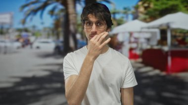 Beyaz tişört giyen ve gözlüklü bir adam, güneş ışığı altında düşünceli bir şekilde çeneye dokunur..