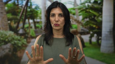 Kadın, geniş yeşil bir parkta, arkasında uzun palmiye ağaçları olan bir geçitte kameraya doğru açık avuçlar tutuyor..