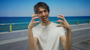 Gözlüklü ve beyaz tişörtlü bir adam mavi gökyüzünün altında okyanus kenarında pençeli elleriyle sırıtıyor..