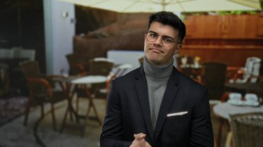 Şehir restoranındaki terasta duran gözlüklü genç adam, dışarıda masa ve şemsiyenin altındaki sandalyelerle yakışıklı ve kendinden emin görünüyor..