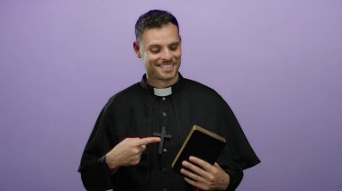 İspanyol rahip İncil 'i işaret ediyor, siyah papaz kıyafeti giyiyor, izole edilmiş mor bir arka plana güvenerek gülümsüyor..