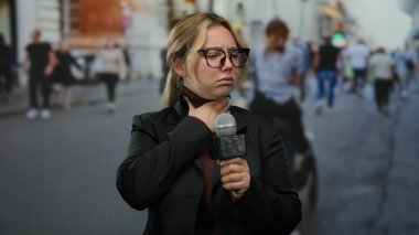 Mikrofon ve gözlüklü kadın muhabir yoğun bir şehir caddesinde arka planda yürüyen insanlarla birlikte dururken boğaz ağrısı çekiyor..