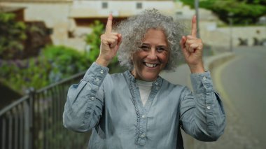 Gri saçlı, gülümseyen ve dışarıyı gösteren bir kadın. Şehir ortamında güler yüzlü, gündüz vakti mavi tişört giyen bir kadın..