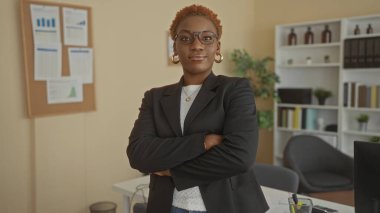 Gözlüklü genç Afrikalı Amerikalı kadın siyah ceketi ilikliyor ve ofis binasında kollarını kavuşturuyor..