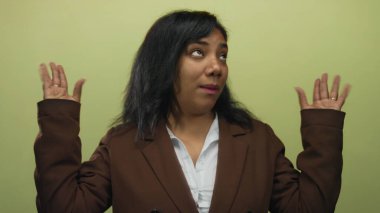 Afrikalı Amerikalı kadın omuz silkiyor ve stüdyoda açık yeşil duvara karşı kahverengi ceket giyen avuç içlerini kaldırıyor..