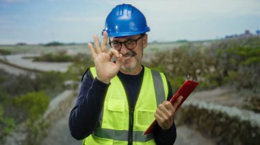 Açık havada, yüksek görüş açısına sahip yelek giyen ve elinde pano olan bir adam, doğal bir manzara arkaplanına gülümseyen, kırsal alanda inşaat ve güvenlik ekipmanlarını gösteren bir adam..