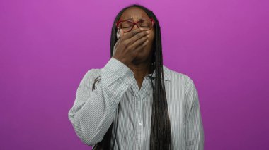 Örgülü ve gözlüklü bir kadın canlı pembe bir arka planda hapşırıyor, ağzını kapatıyor, soğuk bir hissi ifade ediyor..