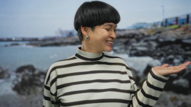 Sahilde gülümseyen bir kadın, açık çizgili bir süveterle okyanus kenarında, kayalık plajlı ve bulutlu gökyüzü arka planda, açık havada, sakin bir şekilde.