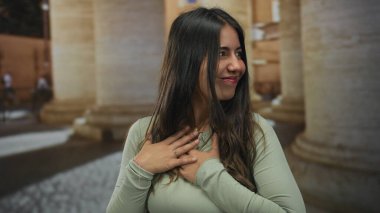 Açık havada gülümseyen, açık yeşil tişörtlü, arka planda büyük sütunlarla rahat bir şekilde duran, sakin ve kendine güvenen bir kadın..