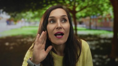 Sarı kazaklı kadın, güneşli park ortamında, yemyeşil ile çevrili, meraklı ve ince yüz hareketleriyle meşgul..