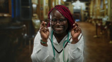 Kapalı bir yerde parmakları çapraz ve steteskopla gülümseyen bir kadın, sıcacık bir kilisede duruyor, ifade yoluyla umut ve huzuru somutlaştırıyor ve ortam yaratıyor..