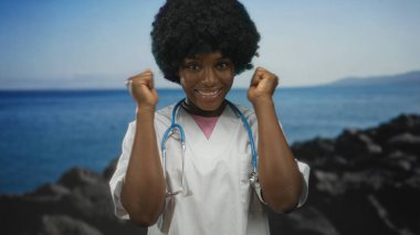 Beyaz önlük ve mavi steteskop giyen genç Afrikalı Amerikalı kadın doktor stüdyoda yumruklarını kaldırdı..