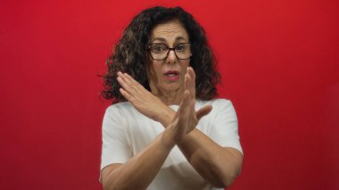 Orta yaşlı İspanyol kadın kırmızı arka plana karşı el kol hareketi yapıyor, reddediyor ya da elleri çapraz, gözlük ve beyaz gömlek giyiyor, kapalı bir yerde izole edilmiş..