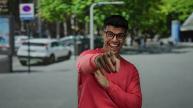 Kırmızı kazaklı genç adam arka planda ağaçlar ve arabalarla güneşli bir caddede kameraya neşeyle gülümsüyor..