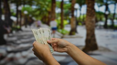 Bir şehir caddesinde elinde Dirham banknotları tutan bir adam ağaçların ve kalabalık bir çevrede yaşayan insanların bulunduğu açık şehir ortamında para birimini sergiliyor..