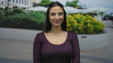 Açık havada duran bir kadın kentin yeşilliği içinde kendine güvenen bir gülümsemeyle ve parlak mor gömleğiyle arka planı bulanık bir şekilde pozitif enerji ve gençlik güvenini yakalıyor..