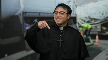 Rahip kılığında gülümseyen bir adam inşaat alanının arka planına işaret ediyor..