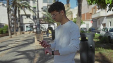 Palmiye ağaçlarının yanındaki güneşli yeşil bir parkta akıllı telefon kullanan ve bileklerine tıklayan bir adam; farkındalık yansıması iç gözlem odağı.