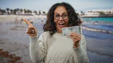 Uae 'de deniz kenarında yaşayan orta yaşlı bir kadın bir dirhem banknotu tutuyor. Şaşırmış ve işaret ediyor. Sahil ve su arka planında canlı bir açık hava sahnesi var..