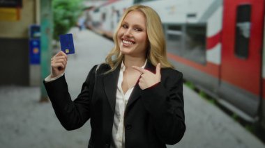 Tren istasyonunda elinde kredi kartıyla gülümseyen, arka planda trenleri olan siyah ceket ve beyaz gömlek giyen bir kadın, seyahat ve finansmanı öneriyor..