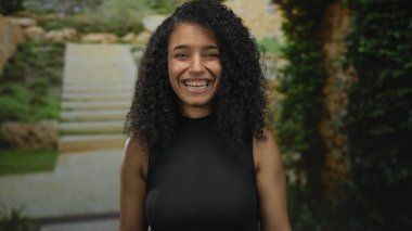 Genç kadın, gözlük takmış parmaklarıyla gözlere çerçeve çiziyor ve sokak basamaklarında kamerayı işaret ediyor; oyuncu merak.