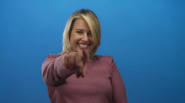 Pembe kazak giyen sarışın kadın stüdyoda kameraya işaret ederken gülüyor ve gülümsüyor..