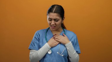 Steteskoplu kadın hemşire göğsünü turuncu bir arka planda rahatsızlık içinde tutuyor. Endişe ve acı hissi taşıyor..