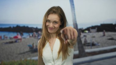 Güneşli sahilde, parlak güneş ışığı altında gülümseyen ve kamerayı işaret eden bir kadın. Dikkat özgüven..
